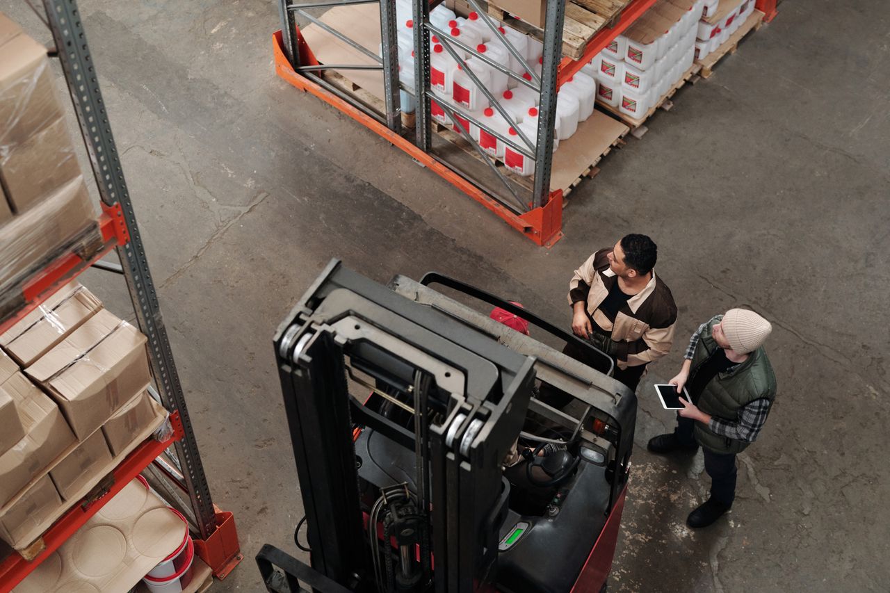 heros-img Two workers in a warehouse discussing logistics near a forklift captured from above.